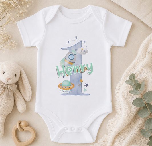 A white onesie with a space-themed design, featuring a rocket ship and stars, is displayed on a bed with a stuffed bunny and a blanket.