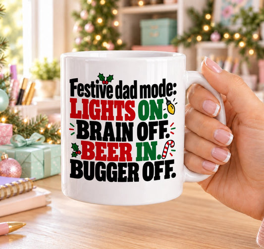 White 11oz ceramic Christmas mug printed with “Festive dad mode: Lights on. Brain off. Beer in. Bugger off.” in red, green and black lettering with a Christmas light bulb, candy cane and holly, held in a festive room