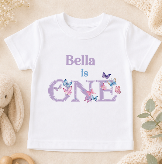 A white t-shirt with the text "Bella is ONE" printed on it, surrounded by colorful butterflies.