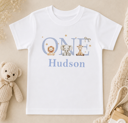 Personalised white t-shirt printed with 'Hudson' and illustrations of a lion, elephant and giraffe, styled with a soft toy and a baby bottle.