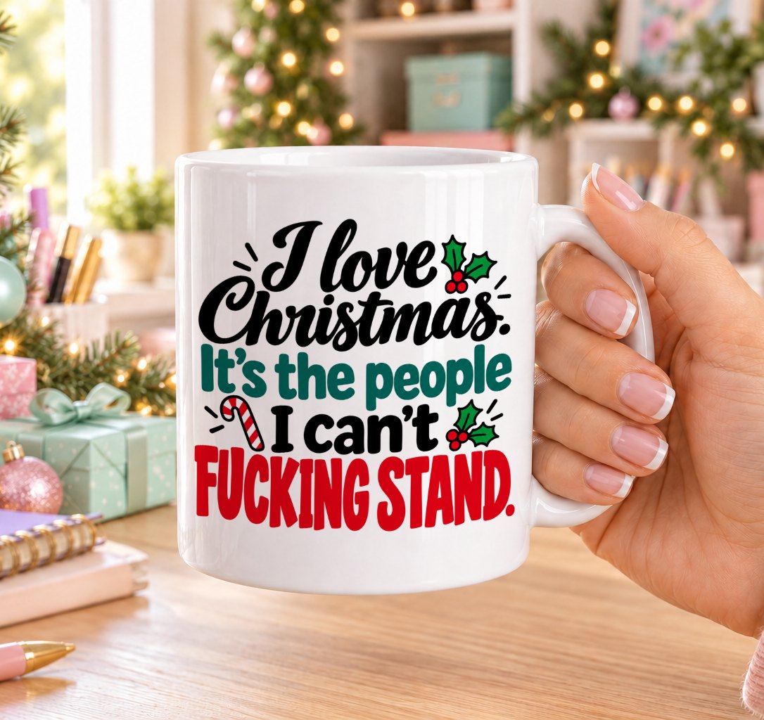 White 11oz ceramic Christmas mug printed with “I love Christmas. It’s the people I can’t f*cking stand.” in red, green and black lettering with a candy cane and holly, held in a festive room