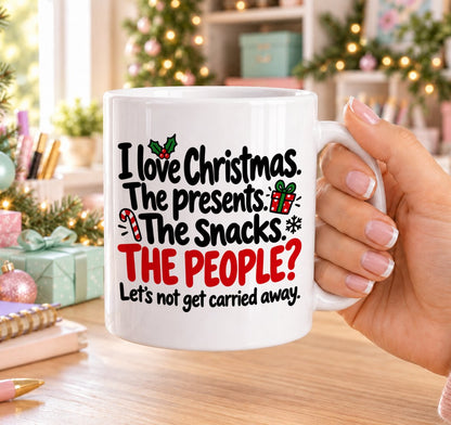White 11oz ceramic Christmas mug printed with “I love Christmas. The presents. The snacks. The people? Let’s not get carried away.” in black and red lettering with a gift box, candy cane, holly and a snowflake, held in a festive room