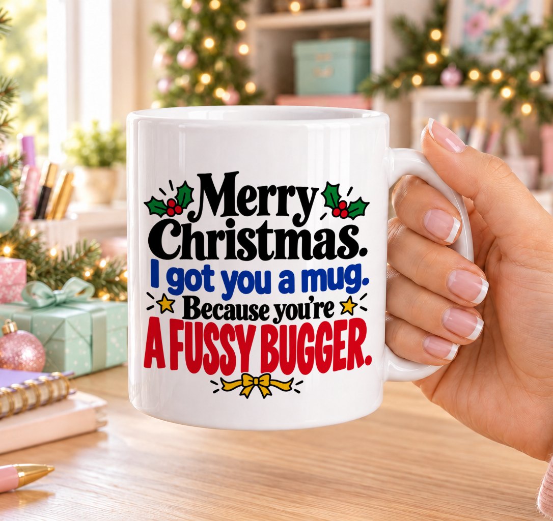 White 11oz ceramic Christmas mug printed with “Merry Christmas. I got you a mug. Because you’re a fussy bugger.” in red, green and black lettering with holly, gold stars and a yellow bow, held in a festive room