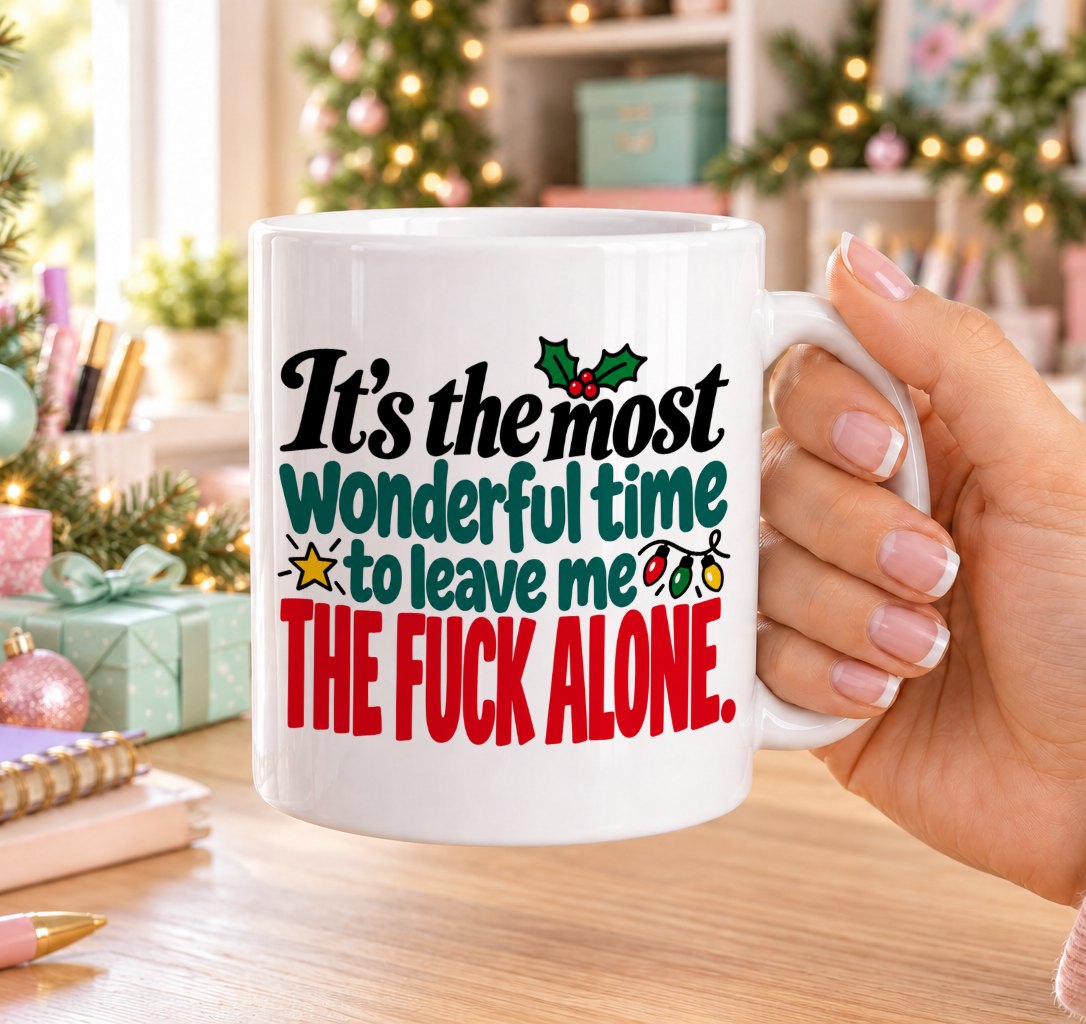 White 11oz ceramic Christmas mug printed with “It’s the most wonderful time to leave me the f*ck alone.” in red, green and black lettering with fairy lights, a gold star and holly, held in a festive room
