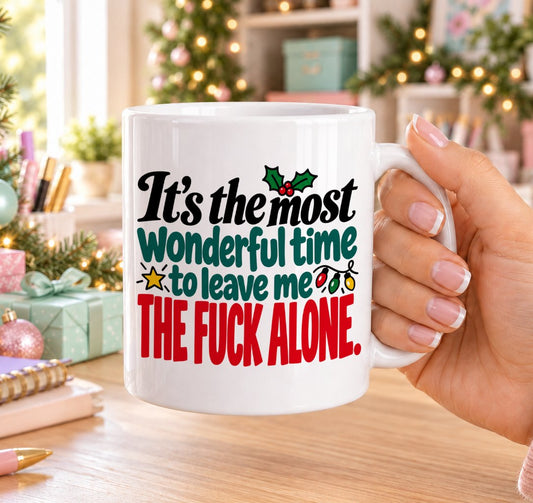White 11oz ceramic Christmas mug printed with “It’s the most wonderful time to leave me the f*ck alone.” in red, green and black lettering with fairy lights, a gold star and holly, held in a festive room