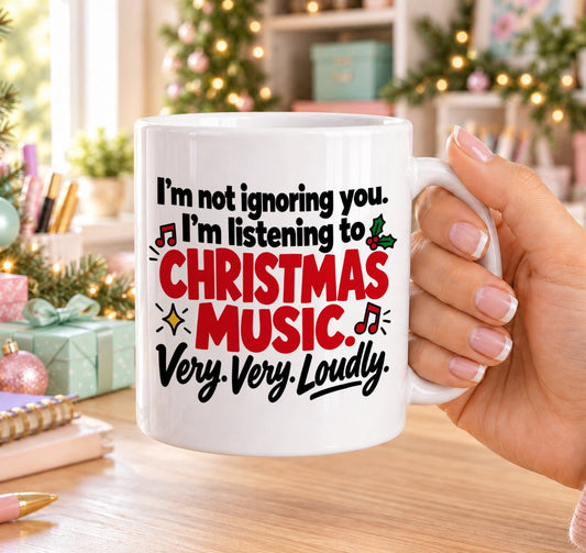 White 11oz ceramic Christmas mug printed with “I’m not ignoring you. I’m listening to Christmas music. Very. Very. Loudly.” in black and red lettering with music notes, holly and gold stars, held in a festive room