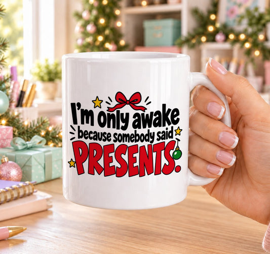White 11oz ceramic Christmas mug printed with “I’m only awake because somebody said presents.” in black and red lettering with a red bow, bauble and gold stars, held in a festive room