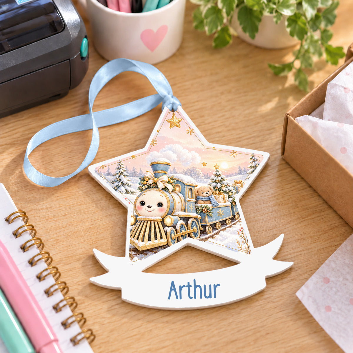 Personalised blue and gold Christmas train white acrylic star bauble with name, 10cm Christmas decoration with ribbon, shown on a desk