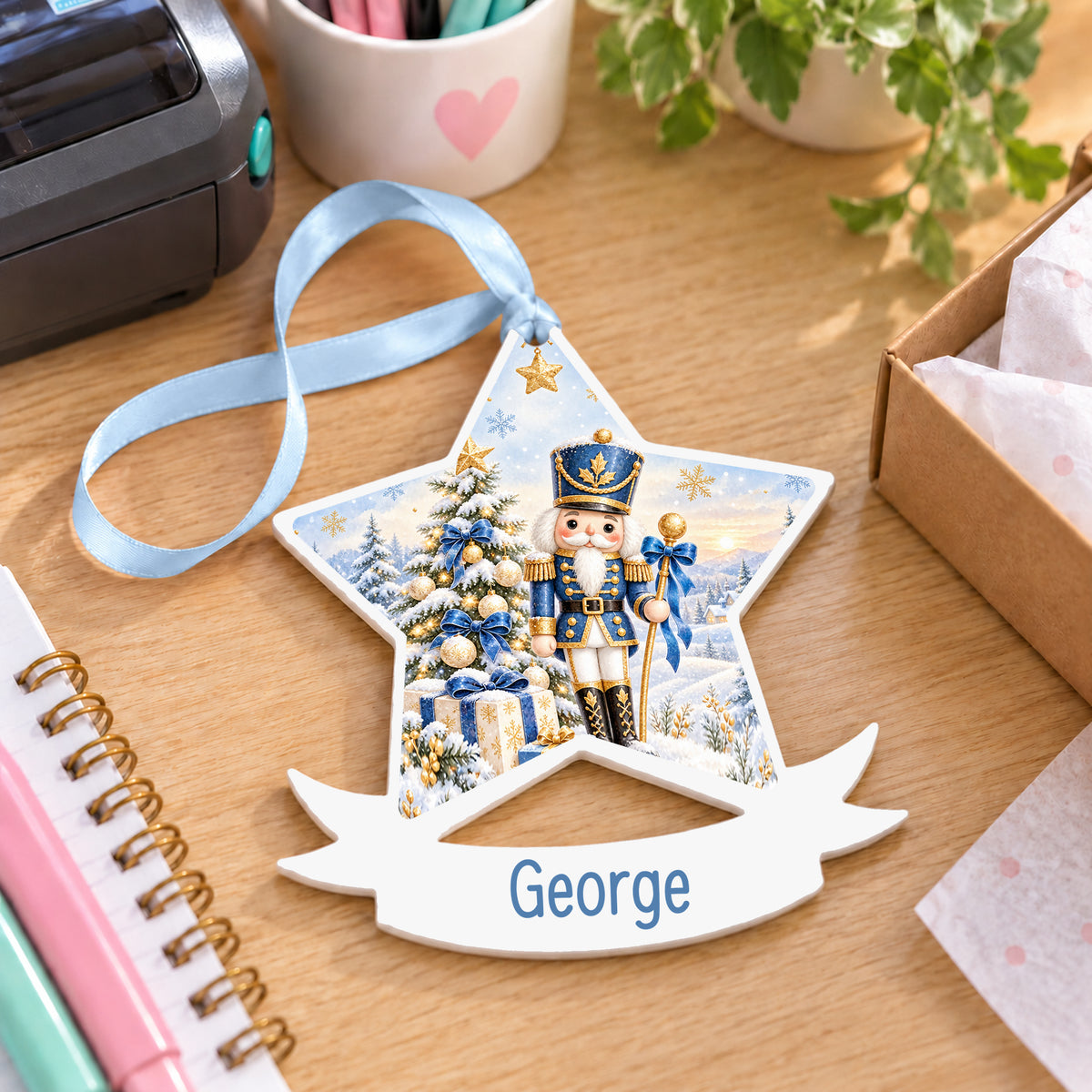 Personalised blue and gold nutcracker white acrylic star bauble with name, 10cm Christmas decoration with ribbon, shown on a desk