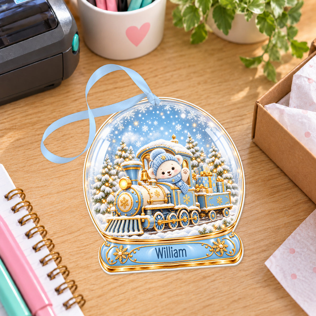 Personalised blue and gold train snow globe bauble with name, white acrylic Christmas decoration with ribbon, shown on a desk