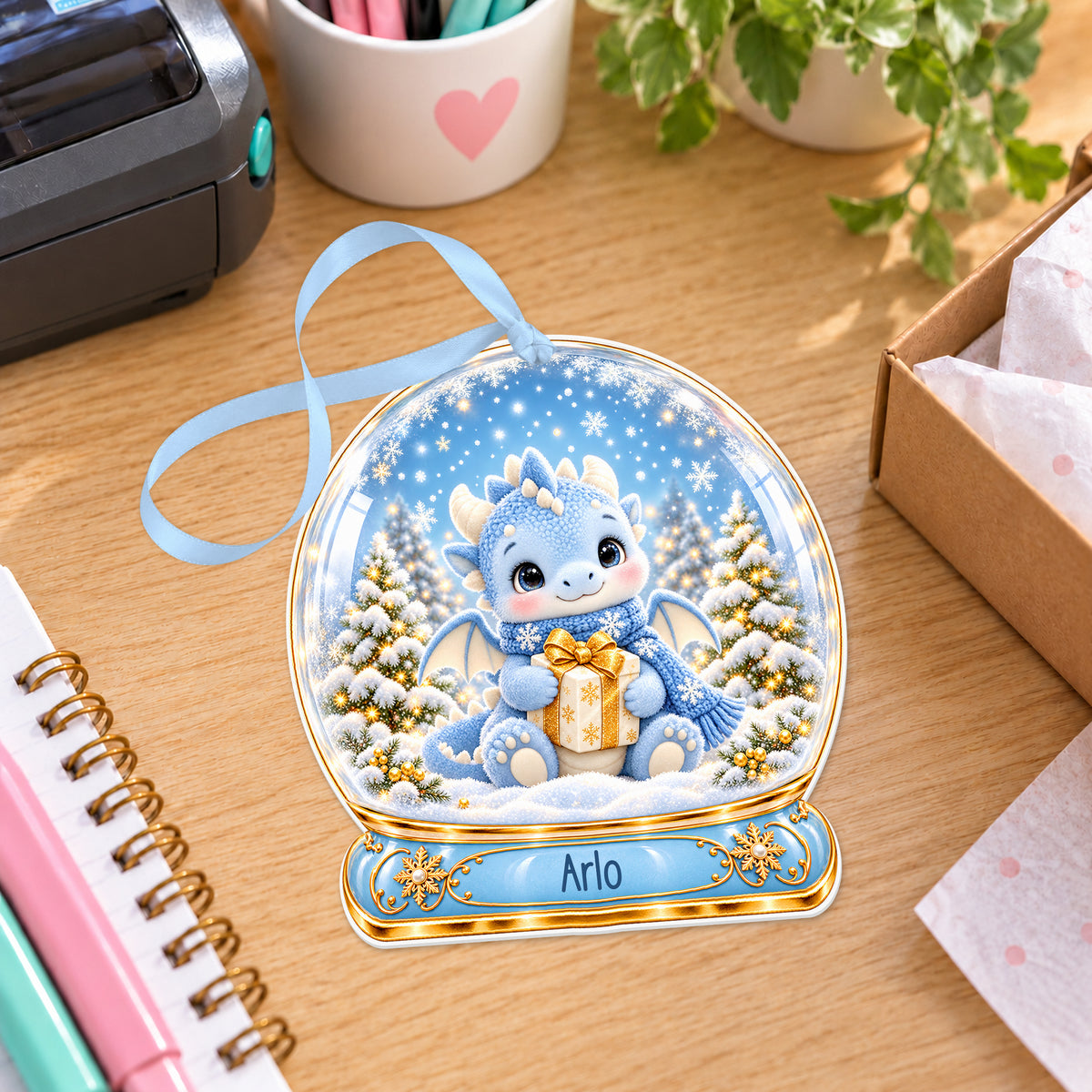 Personalised blue dragon snow globe bauble with name, white acrylic Christmas decoration with ribbon, shown on a desk