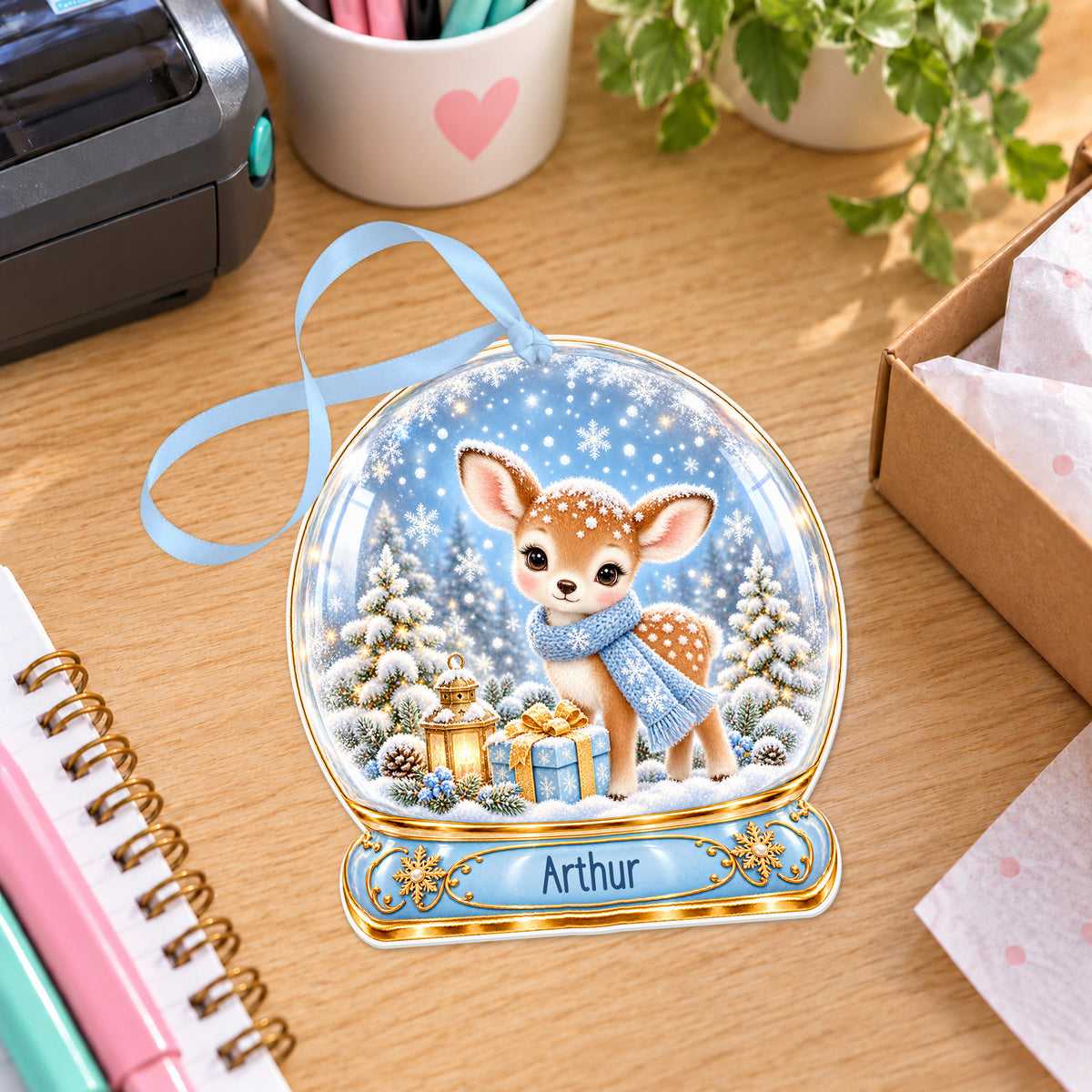 Personalised blue fawn snow globe bauble with name, white acrylic Christmas decoration with ribbon, shown on a desk