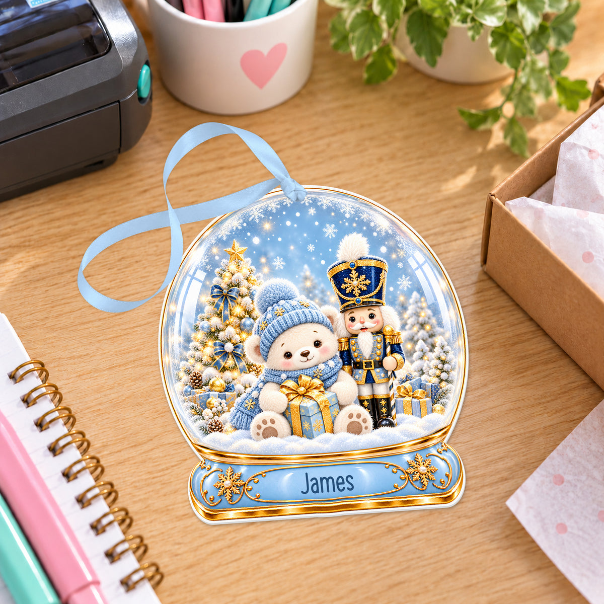 Personalised blue nutcracker snow globe bauble with name, white acrylic Christmas decoration with ribbon, shown on a desk