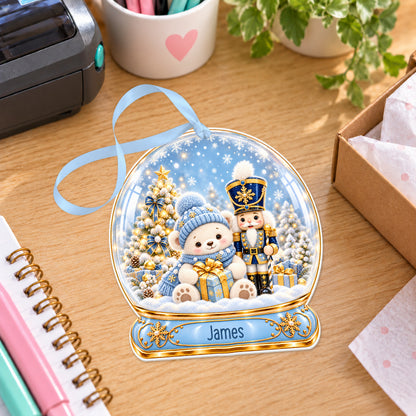 Personalised blue nutcracker snow globe bauble with name, white acrylic Christmas decoration with ribbon, shown on a desk