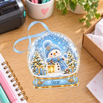 Personalised blue snowman snow globe bauble with name, white acrylic Christmas decoration with ribbon, shown on a desk