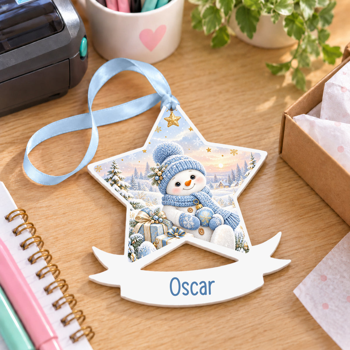 Personalised blue snowman white acrylic star bauble with name, 10cm Christmas decoration with ribbon, shown on a desk