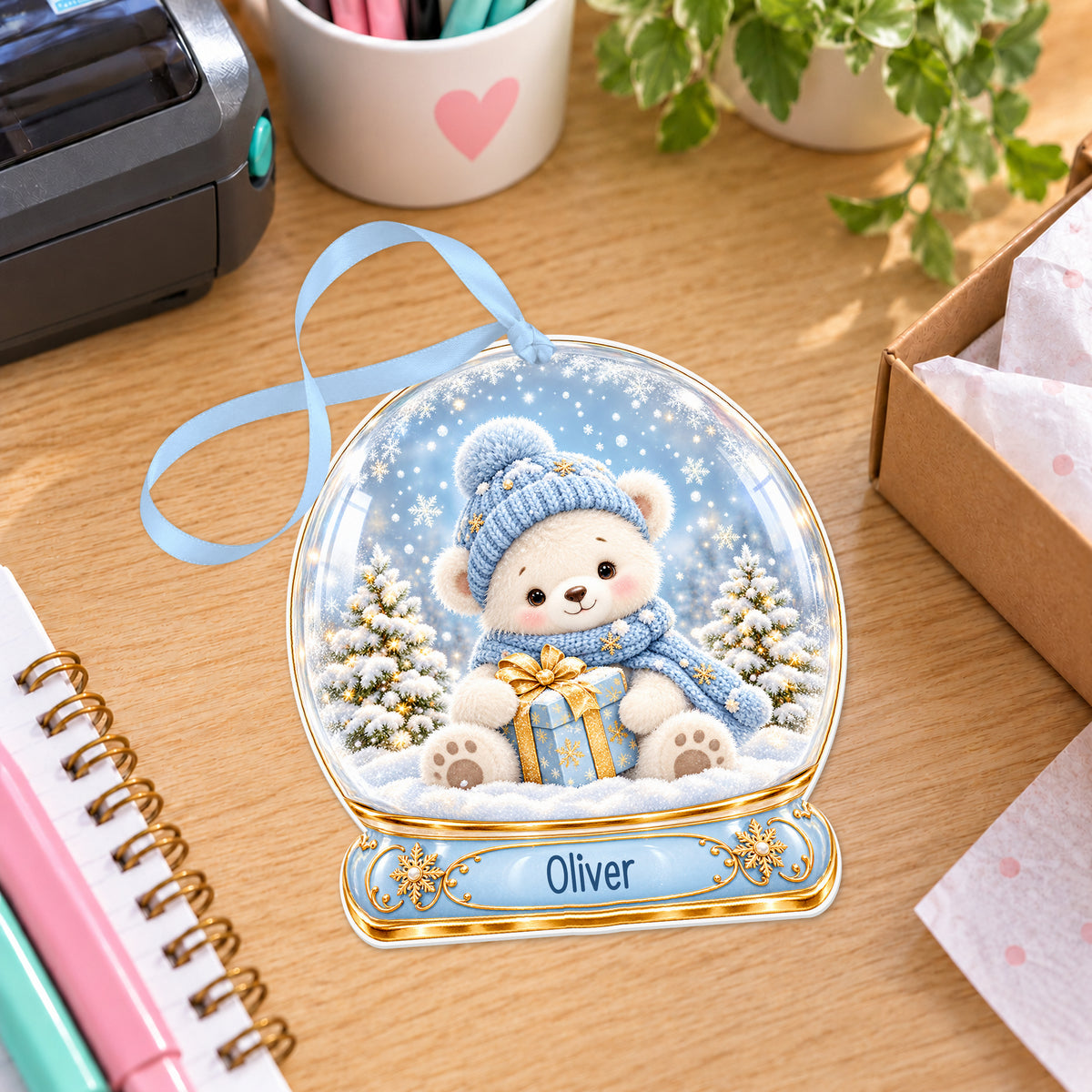 Personalised blue teddy snow globe bauble with name, white acrylic Christmas decoration with ribbon, shown on a desk