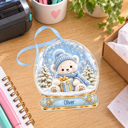 Personalised blue teddy snow globe bauble with name, white acrylic Christmas decoration with ribbon, shown on a desk