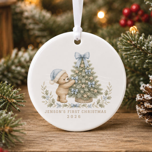 Personalised white ceramic baby’s first Christmas bauble with a teddy bear in a blue Santa hat decorating a tree with blue bows, “Jenson’s First Christmas 2026”
