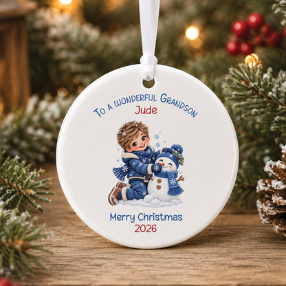 Personalised white ceramic Christmas bauble showing a little boy in a blue snowsuit building a snowman in a matching blue hat and scarf, with To a wonderful Grandson, a name and Merry Christmas 2026