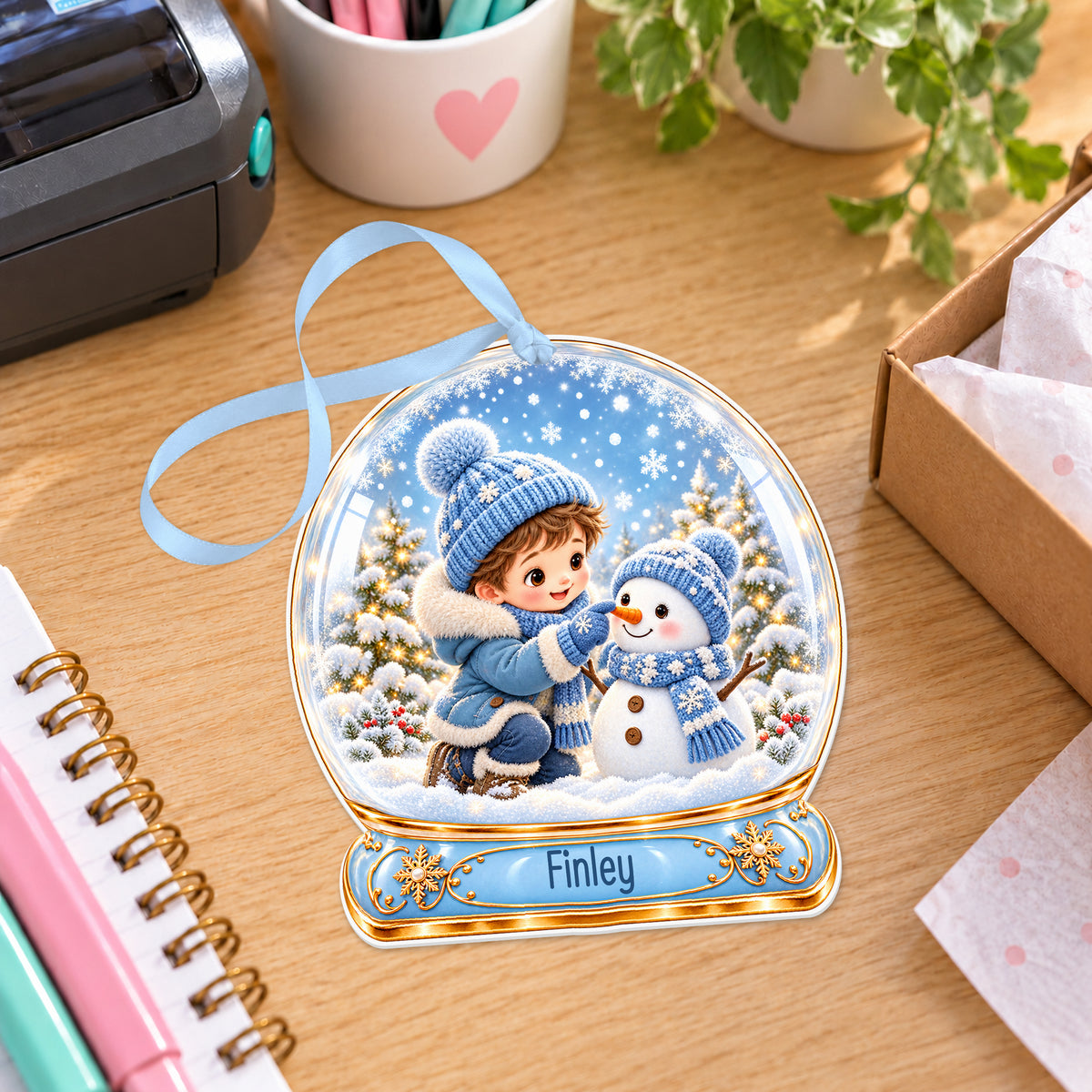 Personalised boy building a snowman snow globe bauble with name, white acrylic Christmas decoration with ribbon, shown on a desk