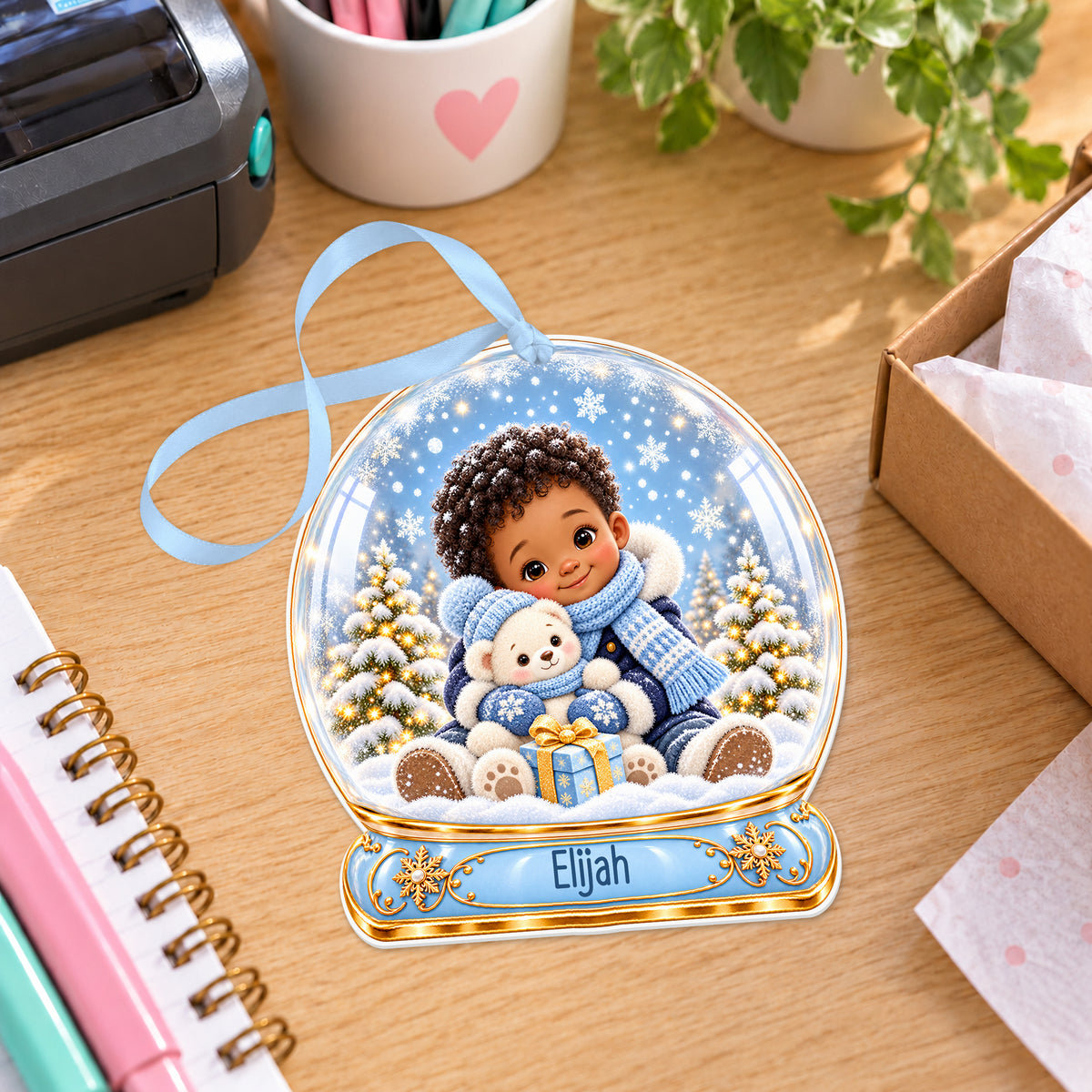 Personalised boy with teddy snow globe bauble with name, white acrylic Christmas decoration with ribbon, shown on a desk
