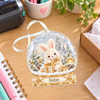 Personalised champagne bunny snow globe bauble with name, white acrylic Christmas decoration with ribbon, shown on a desk