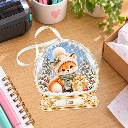 Personalised Christmas fox snow globe bauble with name, white acrylic Christmas decoration with ribbon, shown on a desk