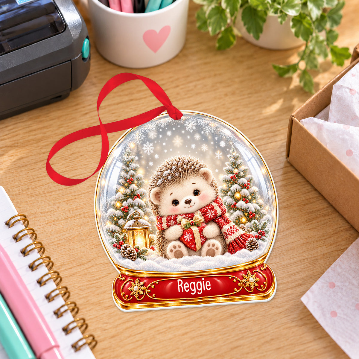 Personalised Christmas hedgehog snow globe bauble with name, white acrylic Christmas decoration with ribbon, shown on a desk