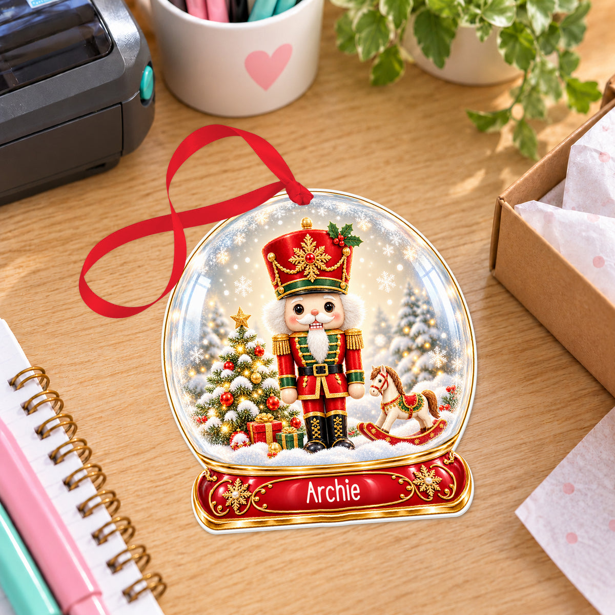 Personalised classic nutcracker snow globe bauble with name, white acrylic Christmas decoration with ribbon, shown on a desk