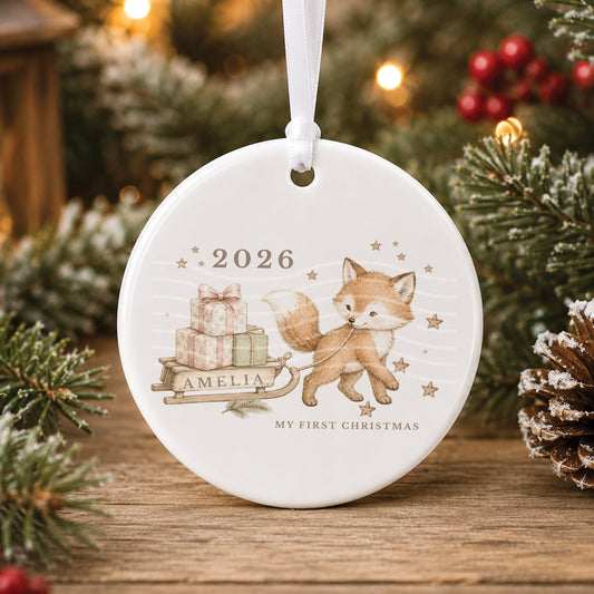 Personalised white ceramic My First Christmas bauble with a little fox pulling a wooden sledge of presents, “Amelia” on the sledge and 2026 among gold stars