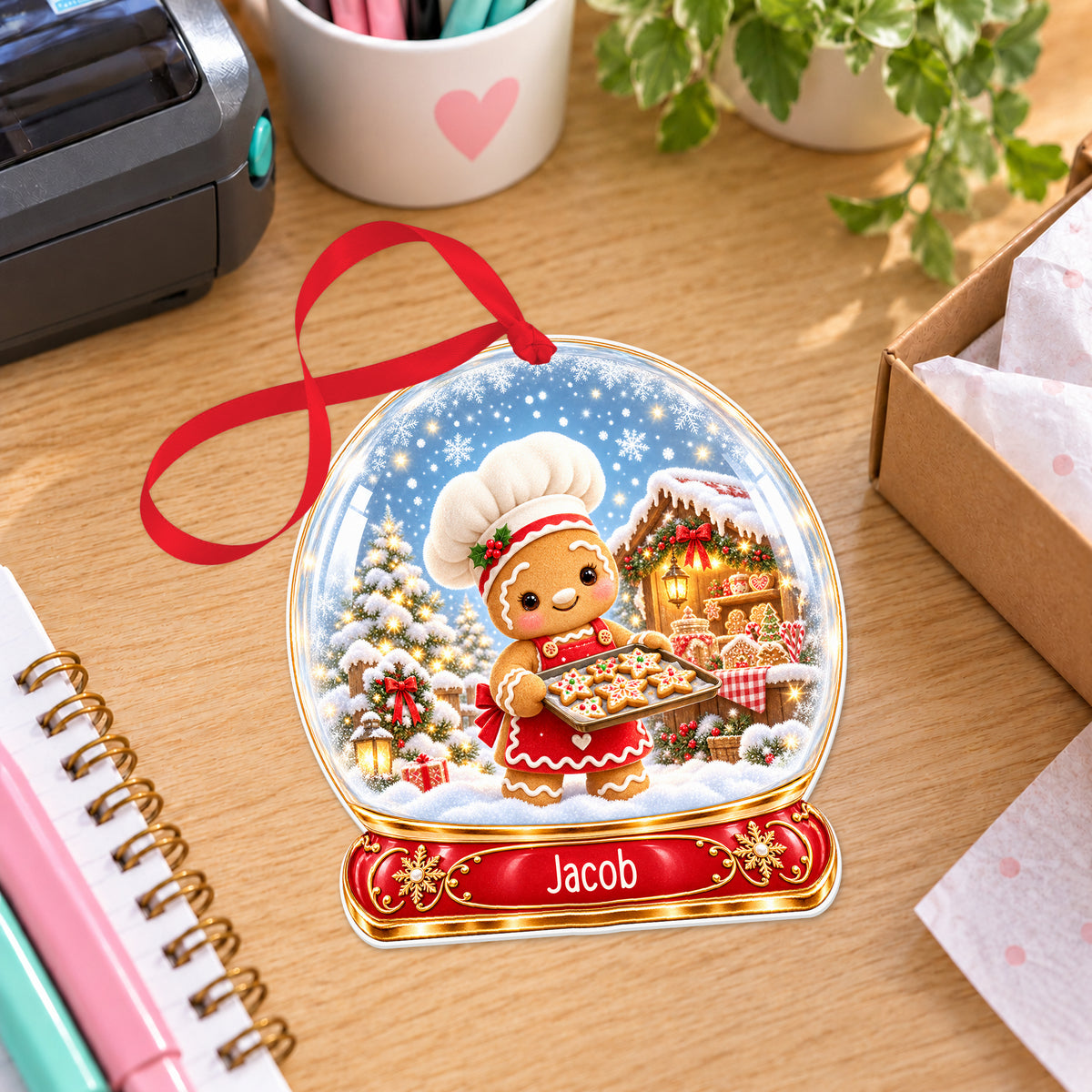 Personalised gingerbread baker snow globe bauble with name, white acrylic Christmas decoration with ribbon, shown on a desk