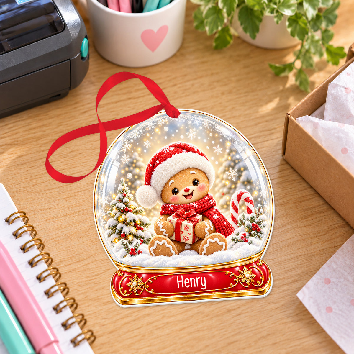 Personalised gingerbread boy snow globe bauble with name, white acrylic Christmas decoration with ribbon, shown on a desk