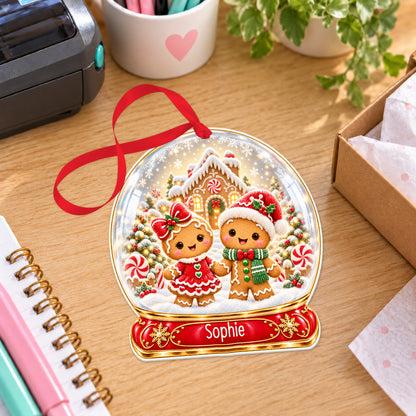 Personalised gingerbread couple snow globe bauble with name, white acrylic Christmas decoration with ribbon, shown on a desk