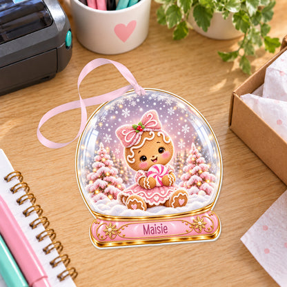 Personalised gingerbread girl snow globe bauble with name, white acrylic Christmas decoration with ribbon, shown on a desk