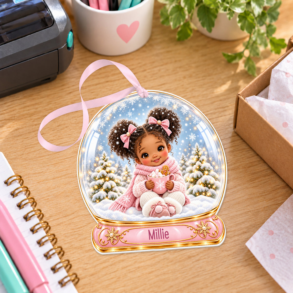 Personalised girl with hot cocoa snow globe bauble with name, white acrylic Christmas decoration with ribbon, shown on a desk