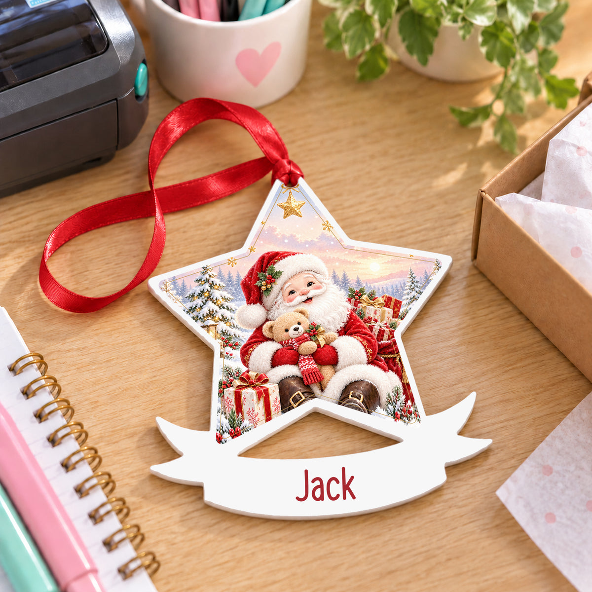 Personalised kind Santa with gifts white acrylic star bauble with name, 10cm Christmas decoration with ribbon, shown on a desk