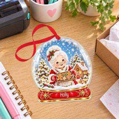 Personalised Mrs Claus baking snow globe bauble with name, white acrylic Christmas decoration with ribbon, shown on a desk