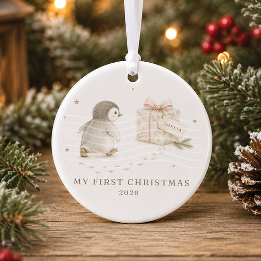 Personalised white ceramic My First Christmas bauble with a baby penguin walking towards a present with a gift tag reading “Amelia”, and 2026