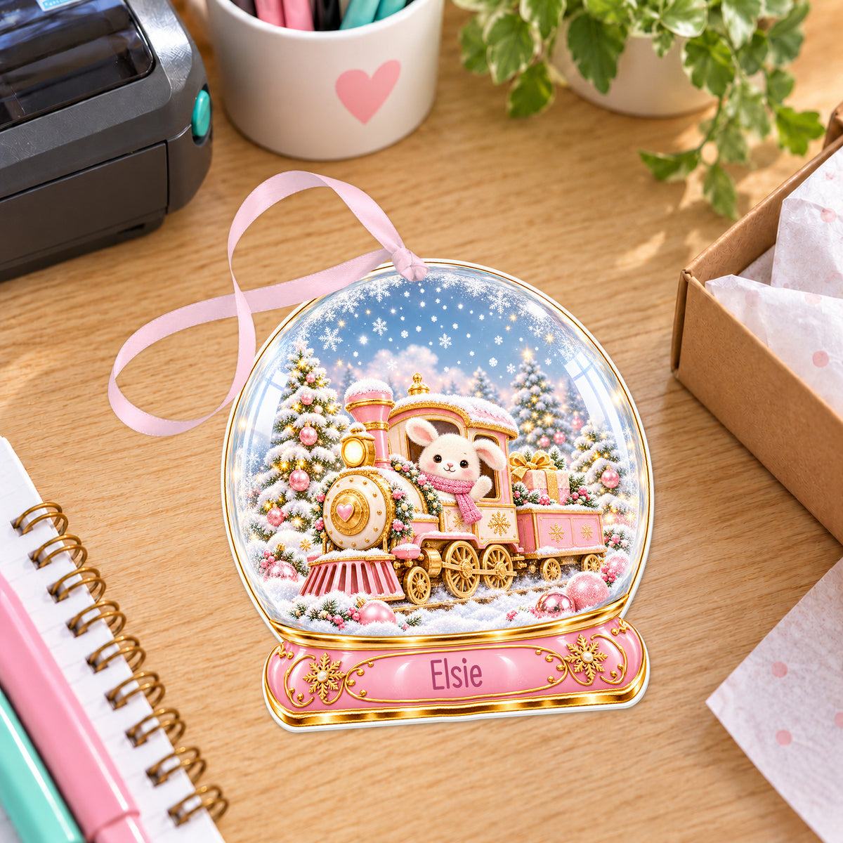 Personalised pink Christmas train snow globe bauble with name, white acrylic Christmas decoration with ribbon, shown on a desk