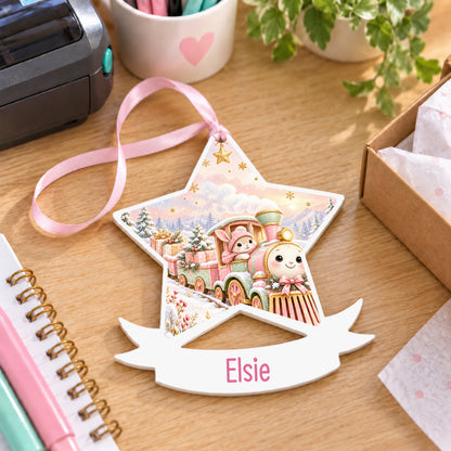 Personalised pink Christmas train white acrylic star bauble with name, 10cm Christmas decoration with ribbon, shown on a desk