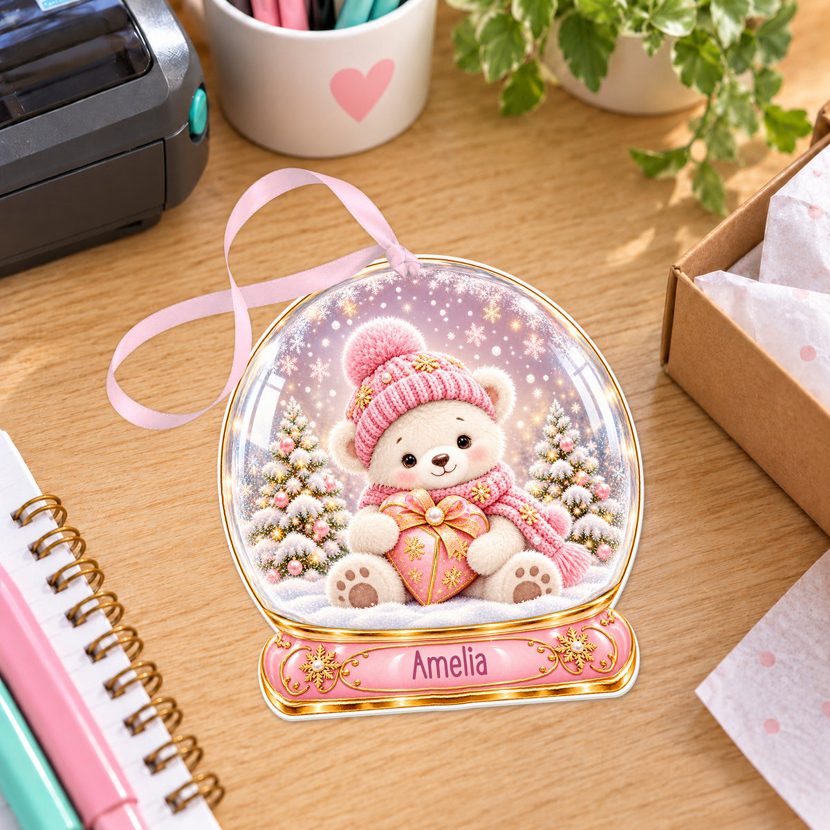 Personalised pink teddy snow globe bauble with name, white acrylic Christmas decoration with ribbon, shown on a desk