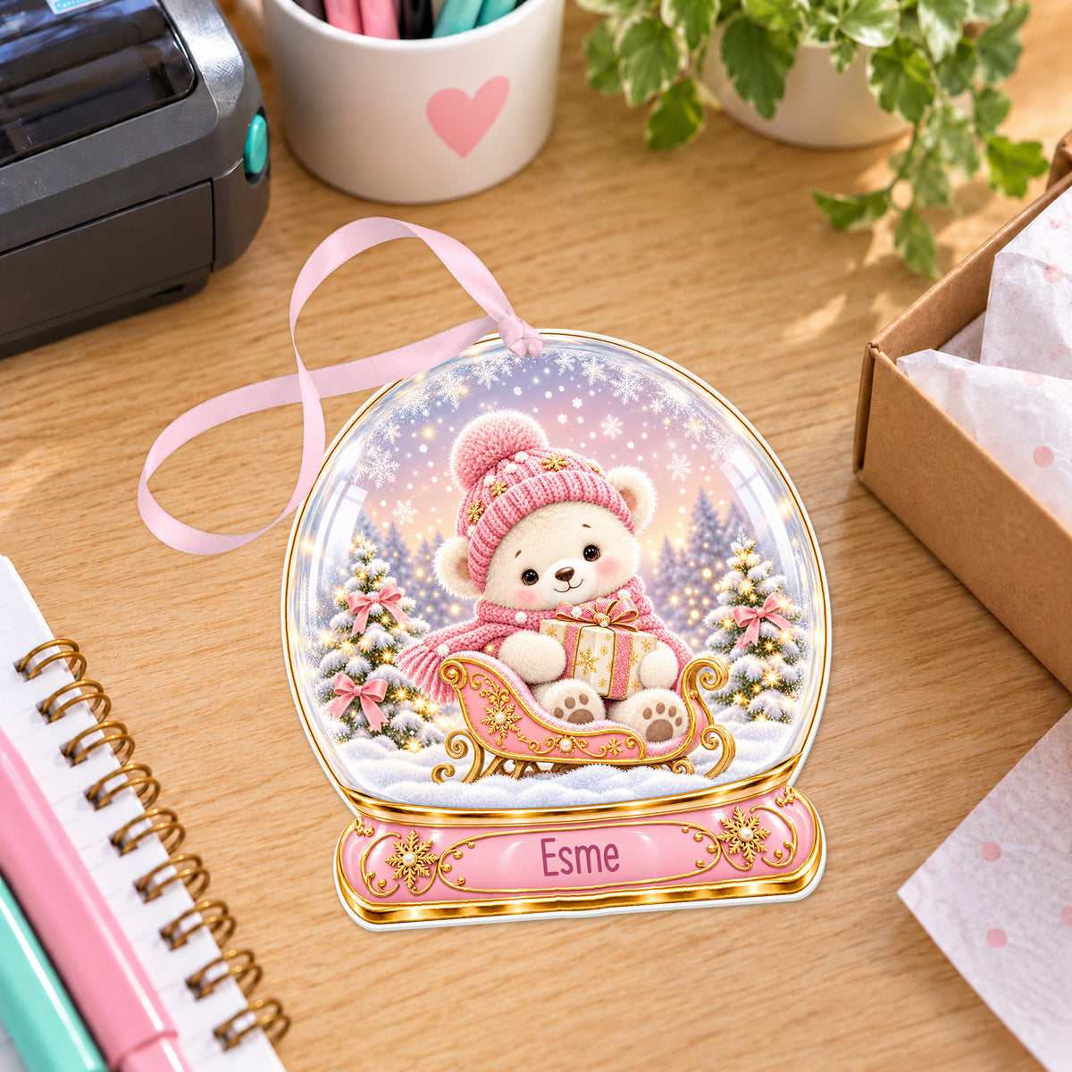Personalised pink teddy sleigh snow globe bauble with name, white acrylic Christmas decoration with ribbon, shown on a desk
