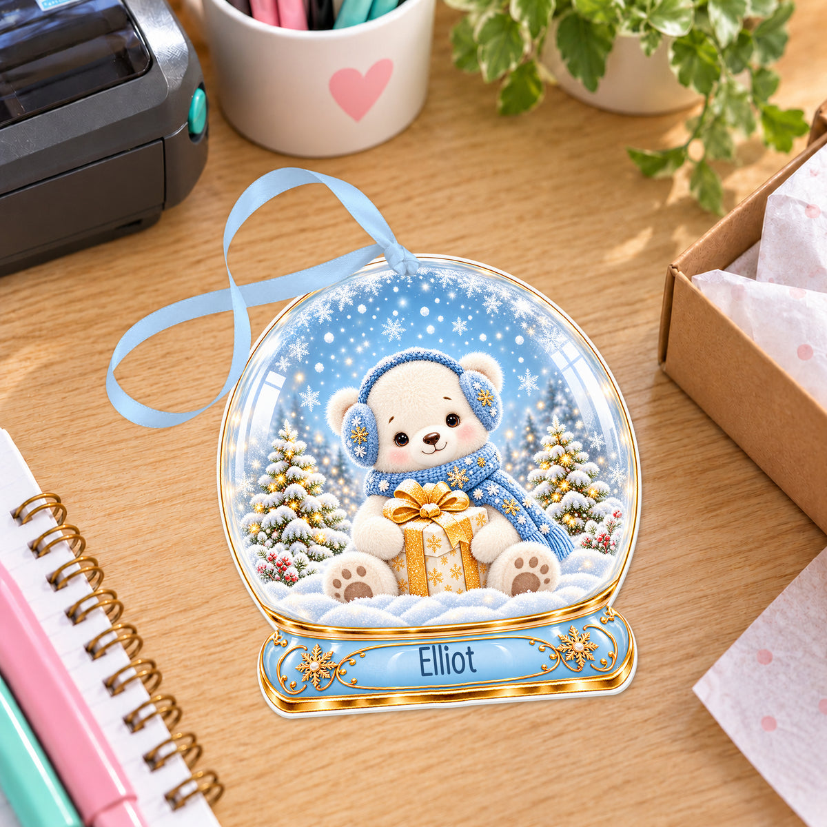 Personalised polar bear cub snow globe bauble with name, white acrylic Christmas decoration with ribbon, shown on a desk
