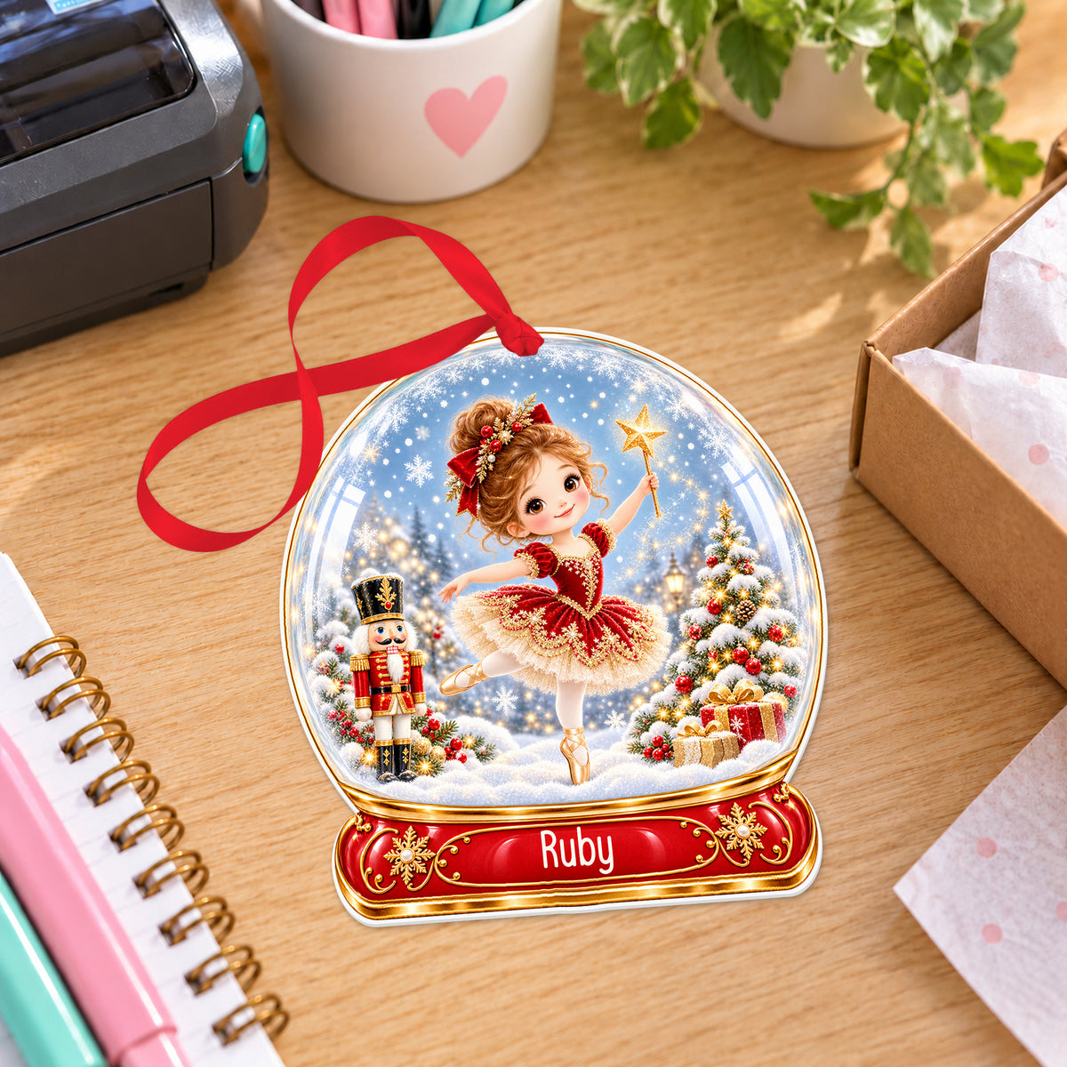 Personalised red ballerina snow globe bauble with name, white acrylic Christmas decoration with ribbon, shown on a desk