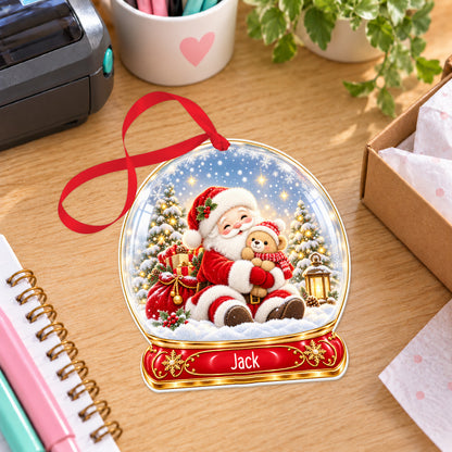 Personalised Santa and teddy snow globe bauble with name, white acrylic Christmas decoration with ribbon, shown on a desk