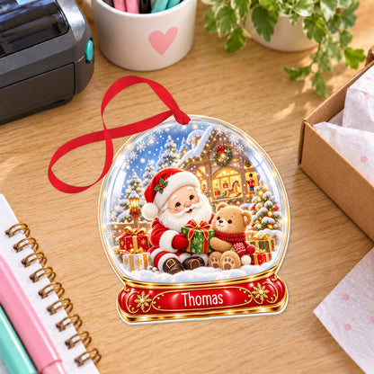 Personalised Santa's workshop snow globe bauble with name, white acrylic Christmas decoration with ribbon, shown on a desk