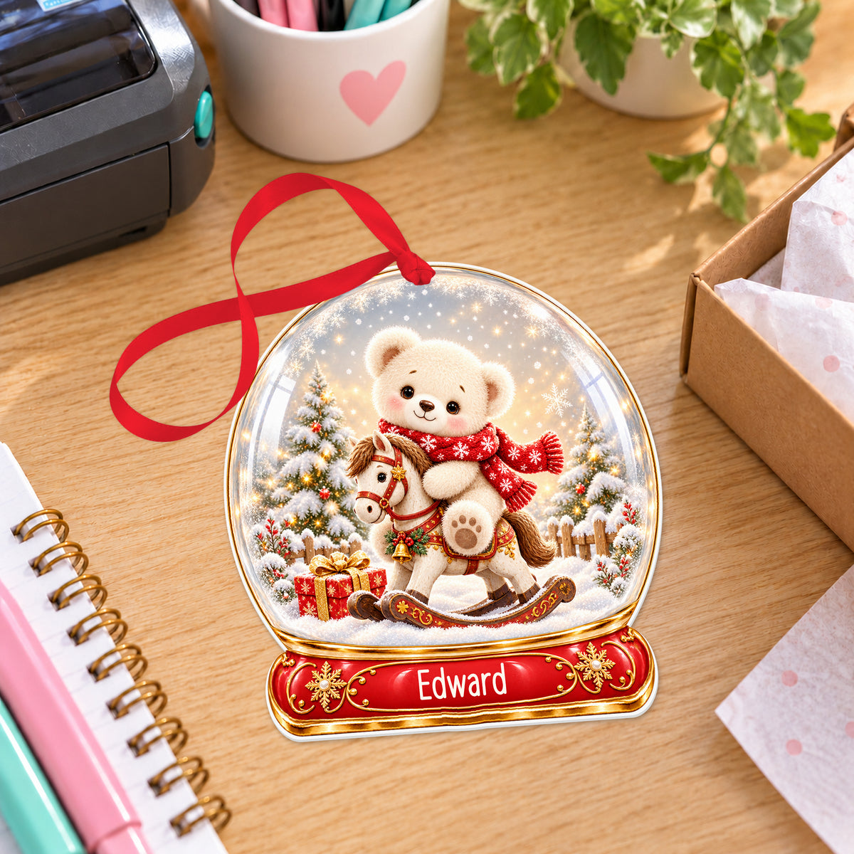 Personalised teddy on rocking horse snow globe bauble with name, white acrylic Christmas decoration with ribbon, shown on a desk