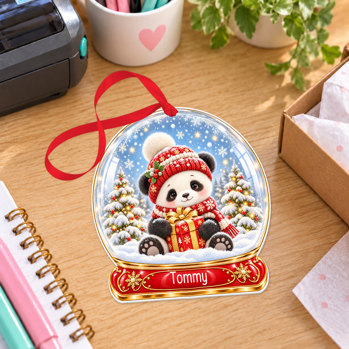 Personalised winter panda snow globe bauble with name, white acrylic Christmas decoration with ribbon, shown on a desk