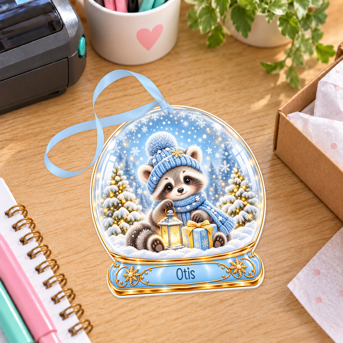 Personalised winter raccoon snow globe bauble with name, white acrylic Christmas decoration with ribbon, shown on a desk